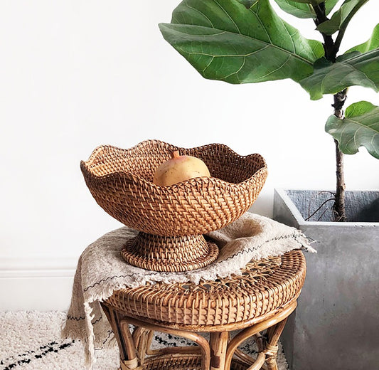 A wicker basket for storage, with a fruit inside, placed on a wicker table in a home setting, with a potted plant in the background.